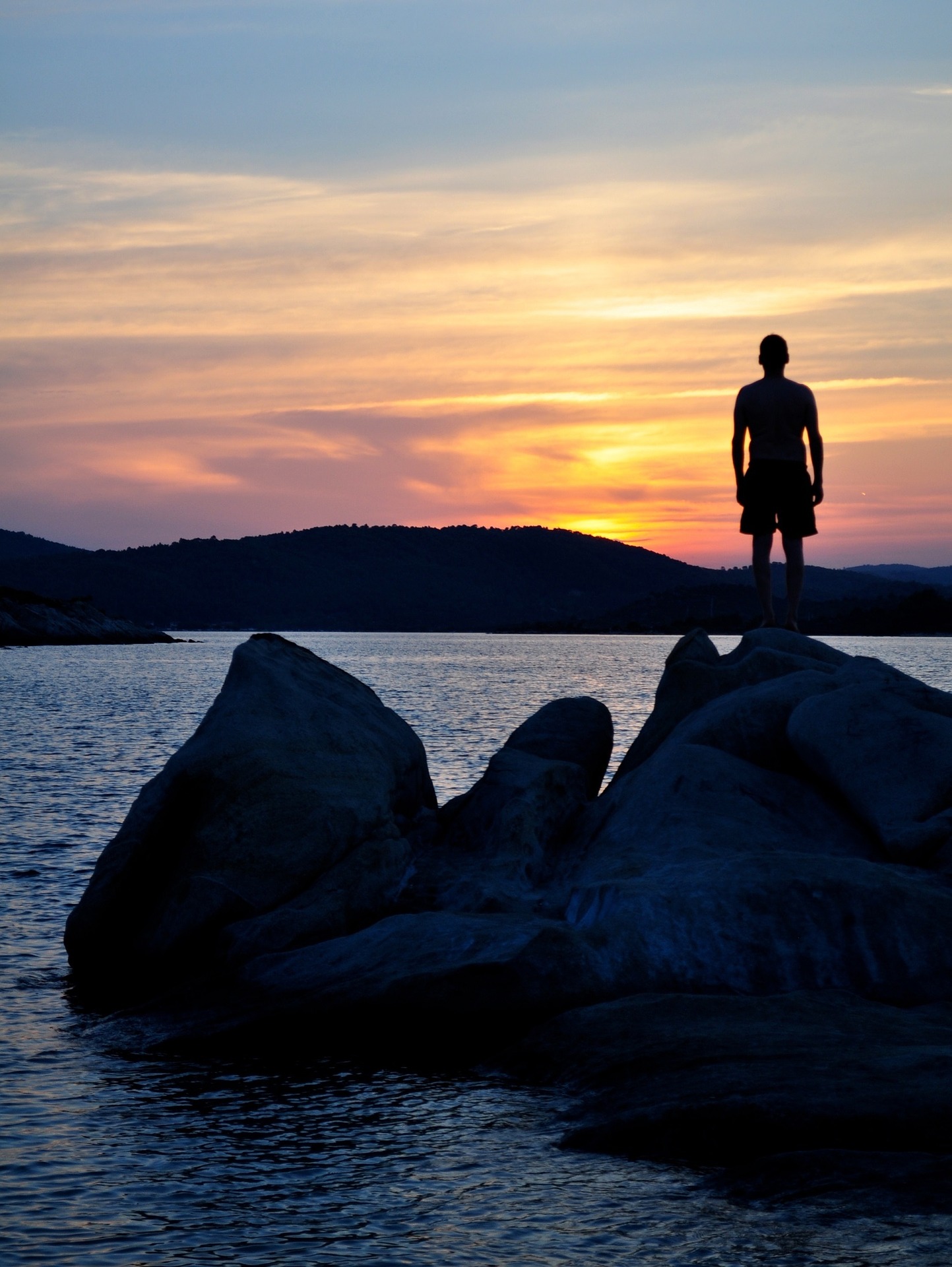 Homem em cima de uma pedra de frente ao mar vendo o pôr-do-sol